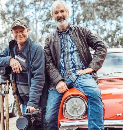 A smiling picture of John Jarratt with his friend sitting on a orange car.